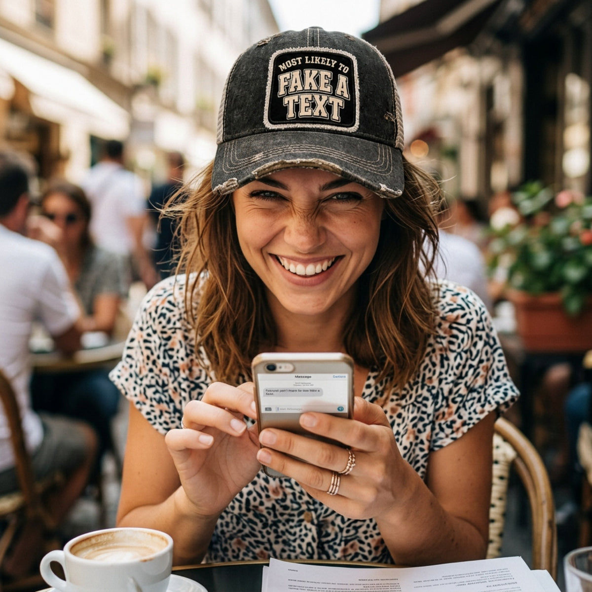 Woman at a sidewalk cafe smiling at her phone, wearing the black MOST LIKELY TO FAKE A TEXT trucker hat