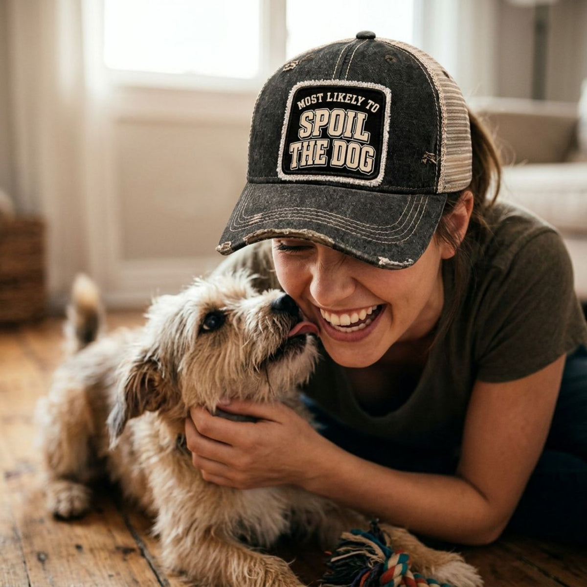 Woman laughing on the floor with her fluffy dog, wearing the black MOST LIKELY TO SPOIL THE DOG trucker hat