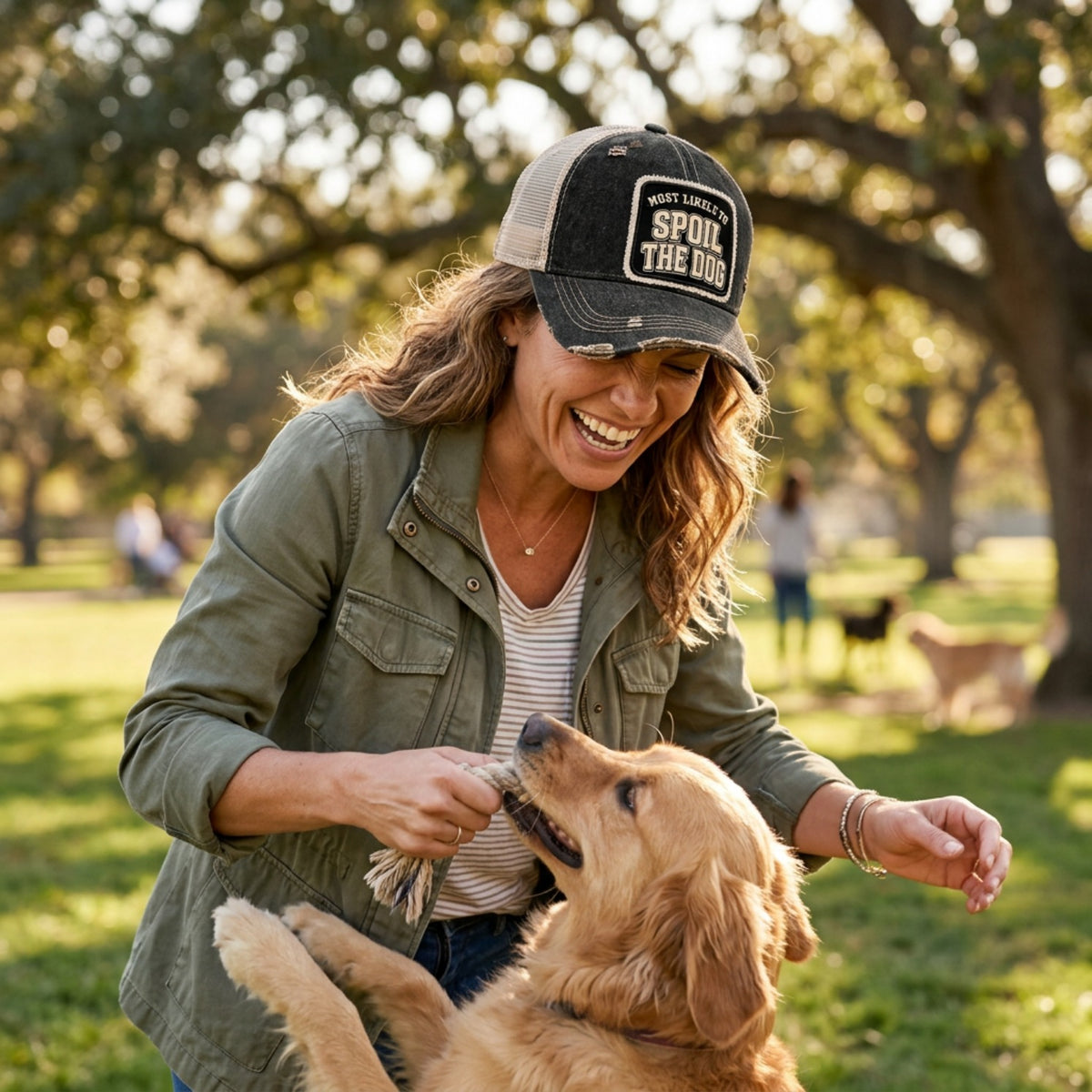 Woman playing with her golden retriever in a park, wearing the black MOST LIKELY TO SPOIL THE DOG trucker hat